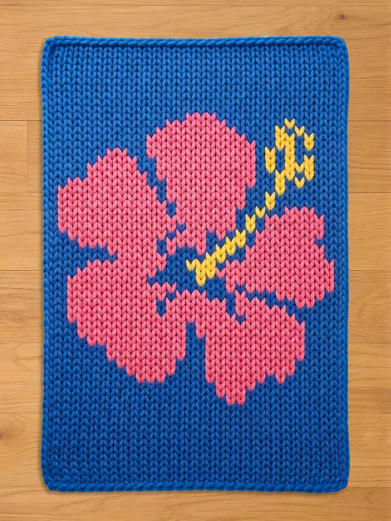 A knitted chunky blanket featuring a pink hibiscus flower on a blue background. The flower's stem and stamen are yellow. The piece is made of thick yarn, creating a textured appearance. The edges are neatly finished.