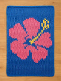 A knitted chunky blanket featuring a pink hibiscus flower on a blue background. The flower's stem and stamen are yellow. The piece is made of thick yarn, creating a textured appearance. The edges are neatly finished.