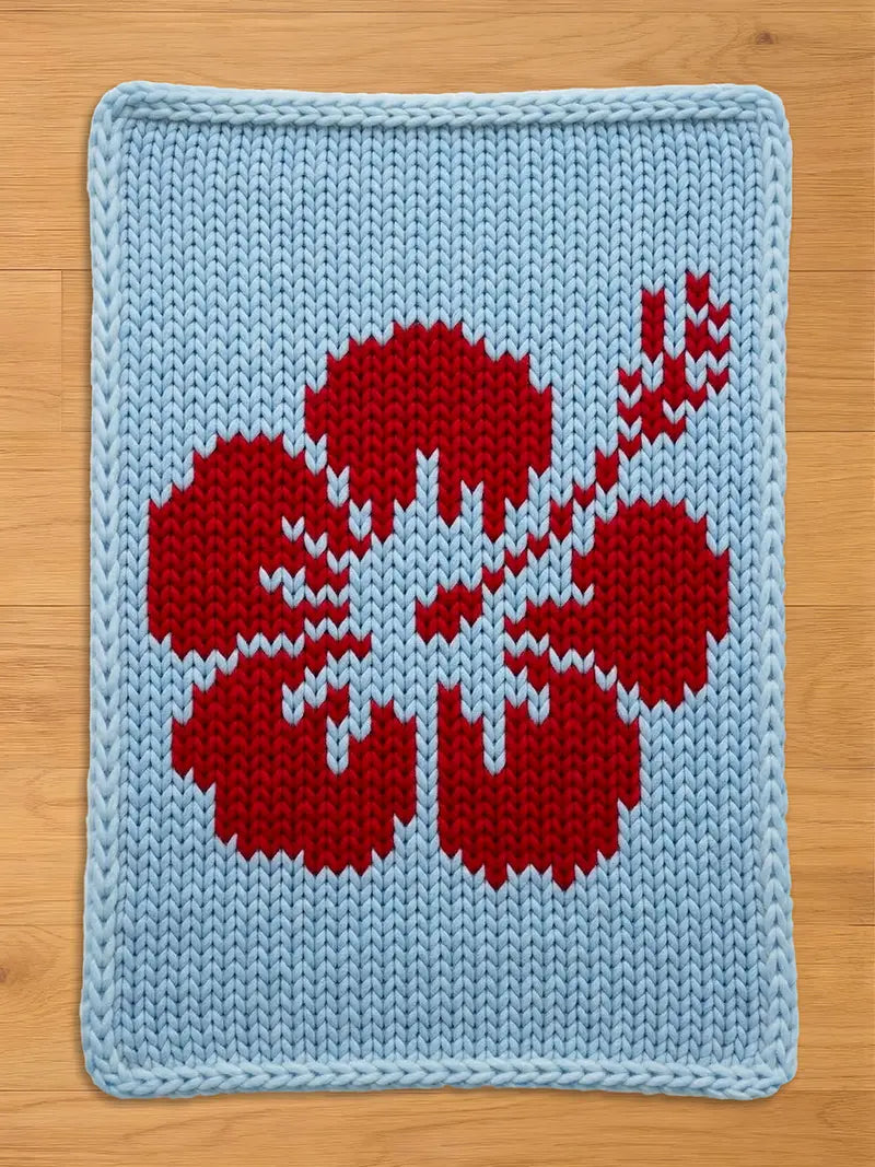 A knitted chunky blanket featuring a red hibiscus flower design on a light blue background. The flower's petals and stem are detailed with white accents. The edges are finished with a light blue border.