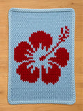 A knitted chunky blanket featuring a red hibiscus flower design on a light blue background. The flower's petals and stem are detailed with white accents. The edges are finished with a light blue border.