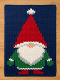 A rectangular knitted textile featuring a gnome character against a dark blue background. The gnome has a tall red pointed hat with a white pom-pom, a large white beard, a peach-colored nose, and a green tunic with red boots.