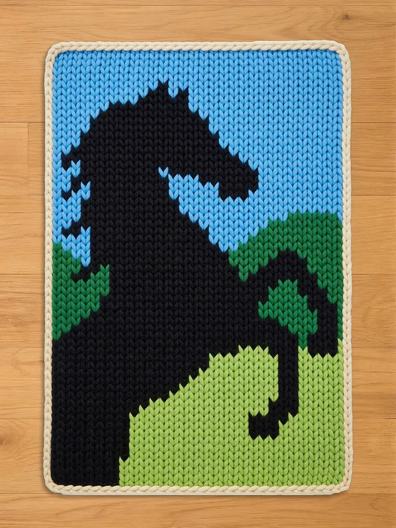 A chunky knit blanket featuring a black rearing horse design against a light blue sky and green hills, finished with a white braided border.
