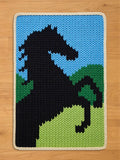A chunky knit blanket featuring a black rearing horse design against a light blue sky and green hills, finished with a white braided border.