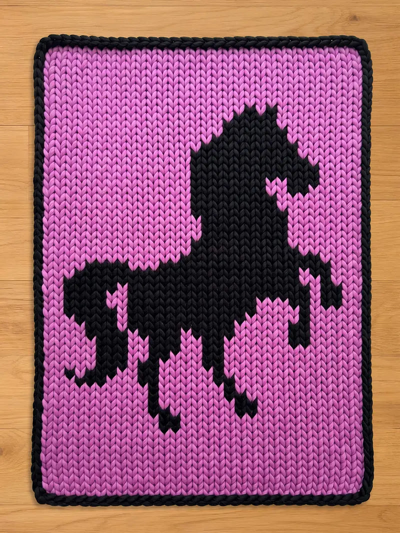 Handmade chunky knit blanket in pink featuring a black prancing horse silhouette design with a black braided border.