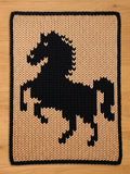 A chunky knit blanket featuring a black rearing horse design on a beige background with a black border, displayed on a wooden surface.
