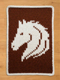Chunky knit blanket featuring a white horse head design woven into a brown background with a white border, displayed on a wooden floor.