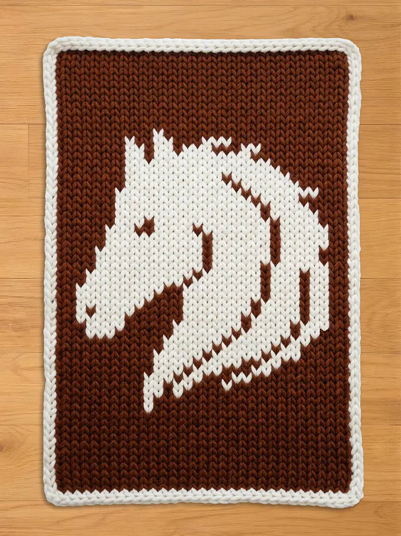 Chunky knit blanket featuring a white horse head design woven into a brown background with a white border, displayed on a wooden floor.
