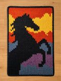 A rectangular, knitted chunky blanket featuring a black horse silhouette against a colorful background. The background transitions from yellow to orange, with accents of blue and purple. The blanket has a black border.