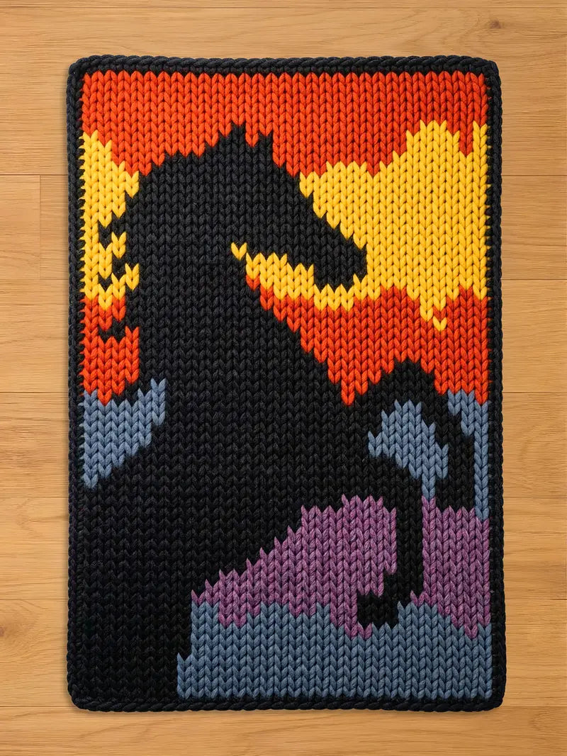 A rectangular, knitted chunky blanket featuring a black horse silhouette against a colorful background. The background transitions from yellow to orange, with accents of blue and purple. The blanket has a black border.