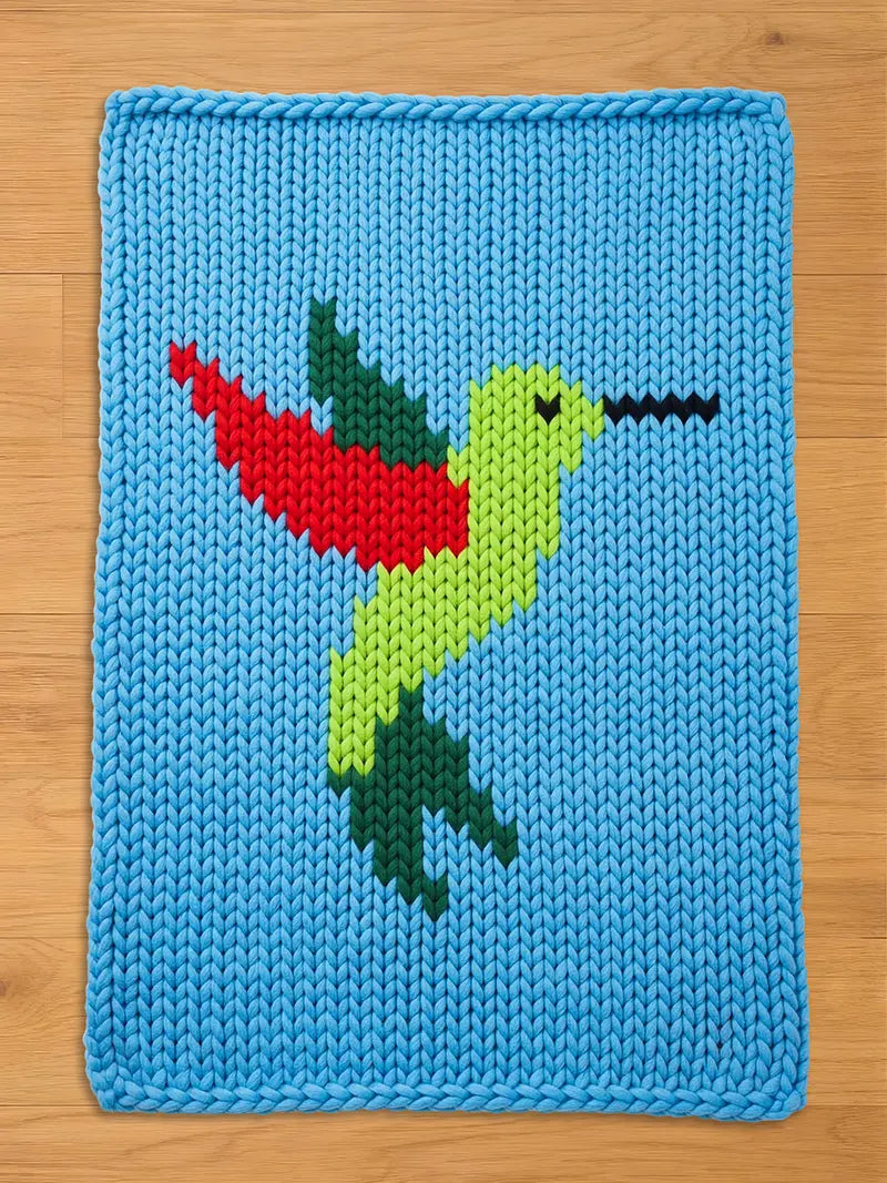 A rectangular chunky knit blanket in light blue, featuring a vibrant hummingbird design knitted into the center. The hummingbird has a bright green body, a red and dark green wing, dark green tail feathers, and a long black beak.