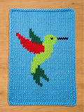 A rectangular chunky knit blanket in light blue, featuring a vibrant hummingbird design knitted into the center. The hummingbird has a bright green body, a red and dark green wing, dark green tail feathers, and a long black beak.