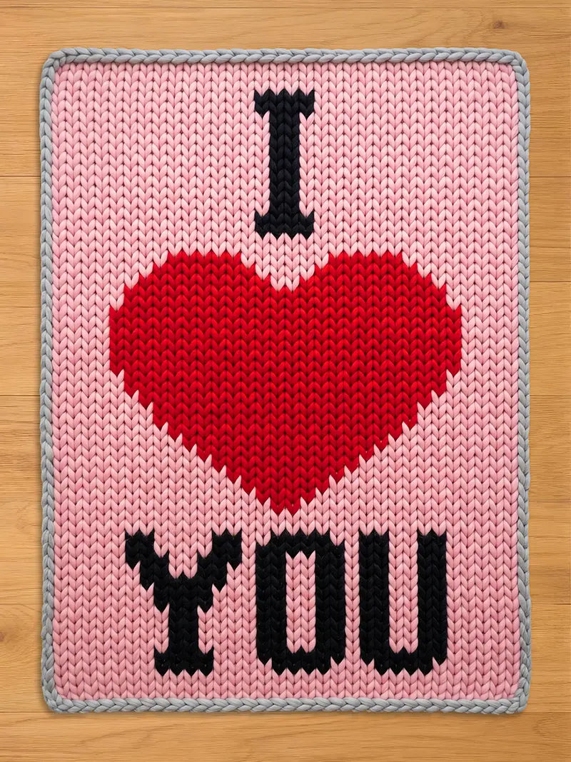 A pink knitted chunky blanket with a red heart and the words "I YOU" in black. The edges are gray. The design is reminiscent of a Valentine's Day theme.