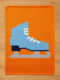 A rectangular chunky knit blanket laid flat on a light wooden surface. The blanket features a vibrant orange background with a large, knitted design of a light blue ice skate in the center, complete with white laces, a dark sole, and a grey blade