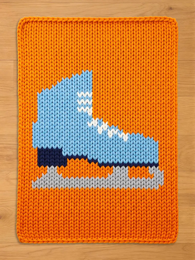 A rectangular chunky knit blanket laid flat on a light wooden surface. The blanket features a vibrant orange background with a large, knitted design of a light blue ice skate in the center, complete with white laces, a dark sole, and a grey blade