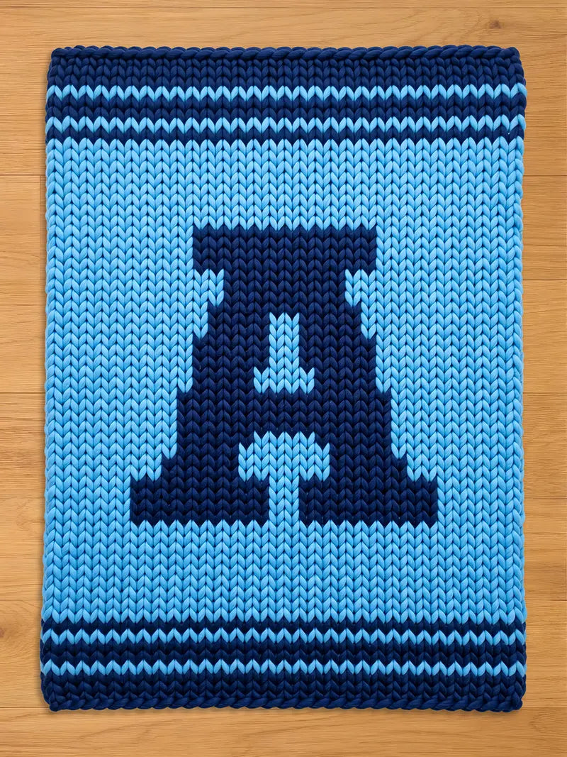  A knitted blanket featuring a large, dark blue letter "A" on a light blue background. The blanket has a textured appearance with a striped border in dark blue. The design is simple and bold, suitable for home decor.