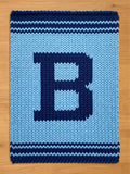 A knitted blanket in shades of blue, featuring a large letter "B" in a darker blue hue. The blanket has a textured appearance with horizontal stripes at the top and bottom, and is displayed on a wooden surface.