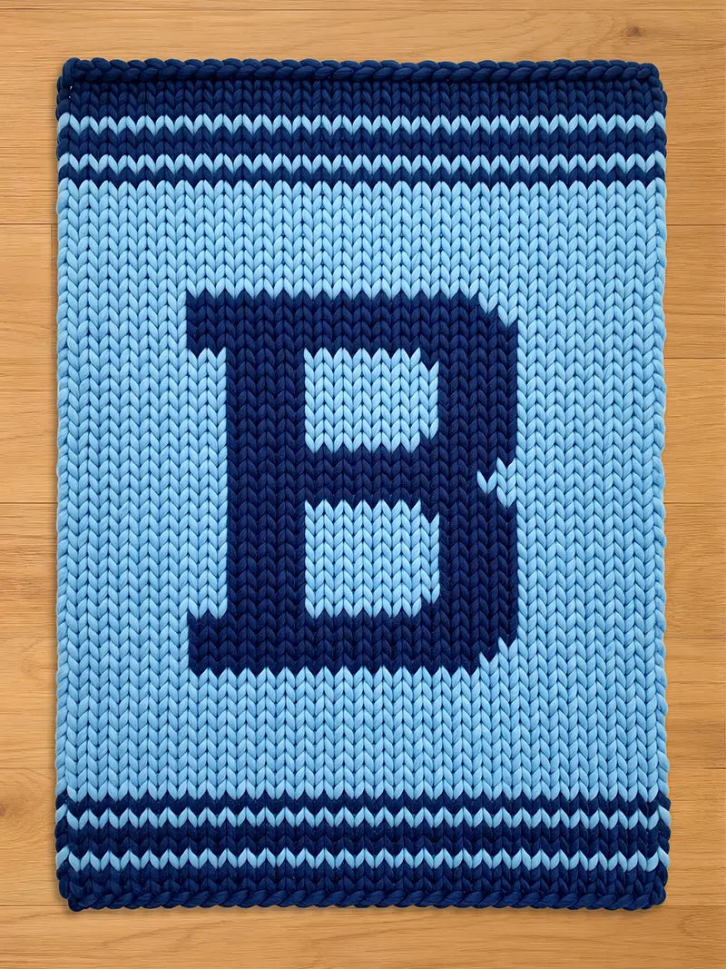 A knitted blanket in shades of blue, featuring a large letter "B" in a darker blue hue. The blanket has a textured appearance with horizontal stripes at the top and bottom, and is displayed on a wooden surface.