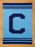 A knitted blanket in shades of blue, featuring a large letter "C" in navy blue against a lighter blue background. The blanket has navy blue stripes at the top and bottom.