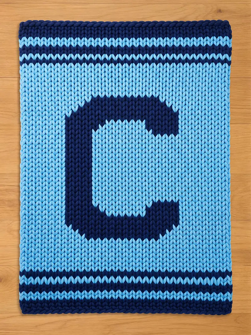 A knitted blanket in shades of blue, featuring a large letter "C" in navy blue against a lighter blue background. The blanket has navy blue stripes at the top and bottom.
