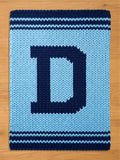 A knitted blanket in light blue with a large navy blue letter "D" in the center. The blanket has navy blue stripes at the top and bottom edges, creating a bold, graphic design. The chunky knit texture is visible.