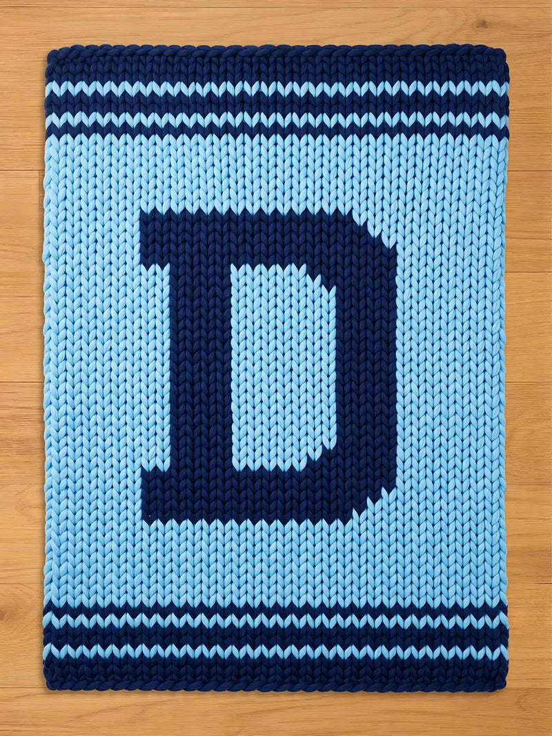 A knitted blanket in light blue with a large navy blue letter "D" in the center. The blanket has navy blue stripes at the top and bottom edges, creating a bold, graphic design. The chunky knit texture is visible.