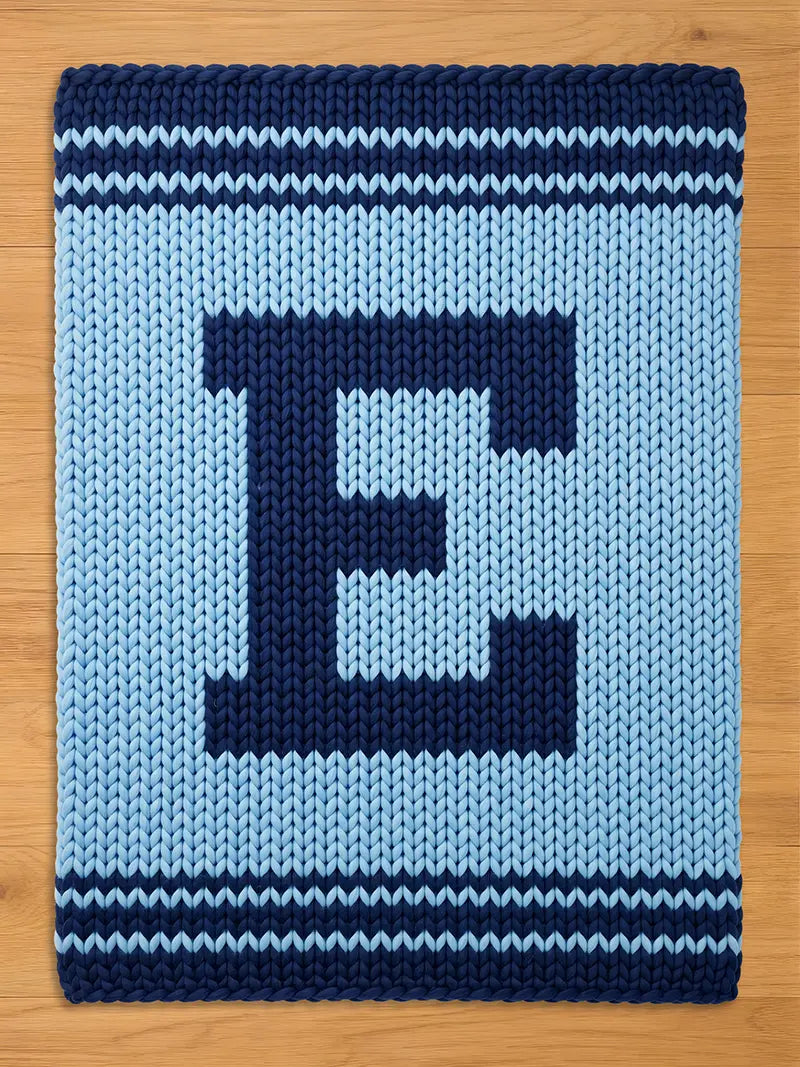 A knitted blanket in shades of blue, featuring a large letter "E" in navy blue against a light blue background. The blanket has navy blue stripes along the top and bottom edges, creating a decorative border.