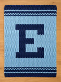A knitted blanket in shades of blue, featuring a large letter "E" in navy blue against a light blue background. The blanket has navy blue stripes along the top and bottom edges, creating a decorative border.