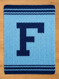 A knitted blanket in shades of blue, featuring a large, dark blue letter "F" in the center. The blanket has a light blue background with dark blue stripes at the top and bottom. The blanket is rectangular.