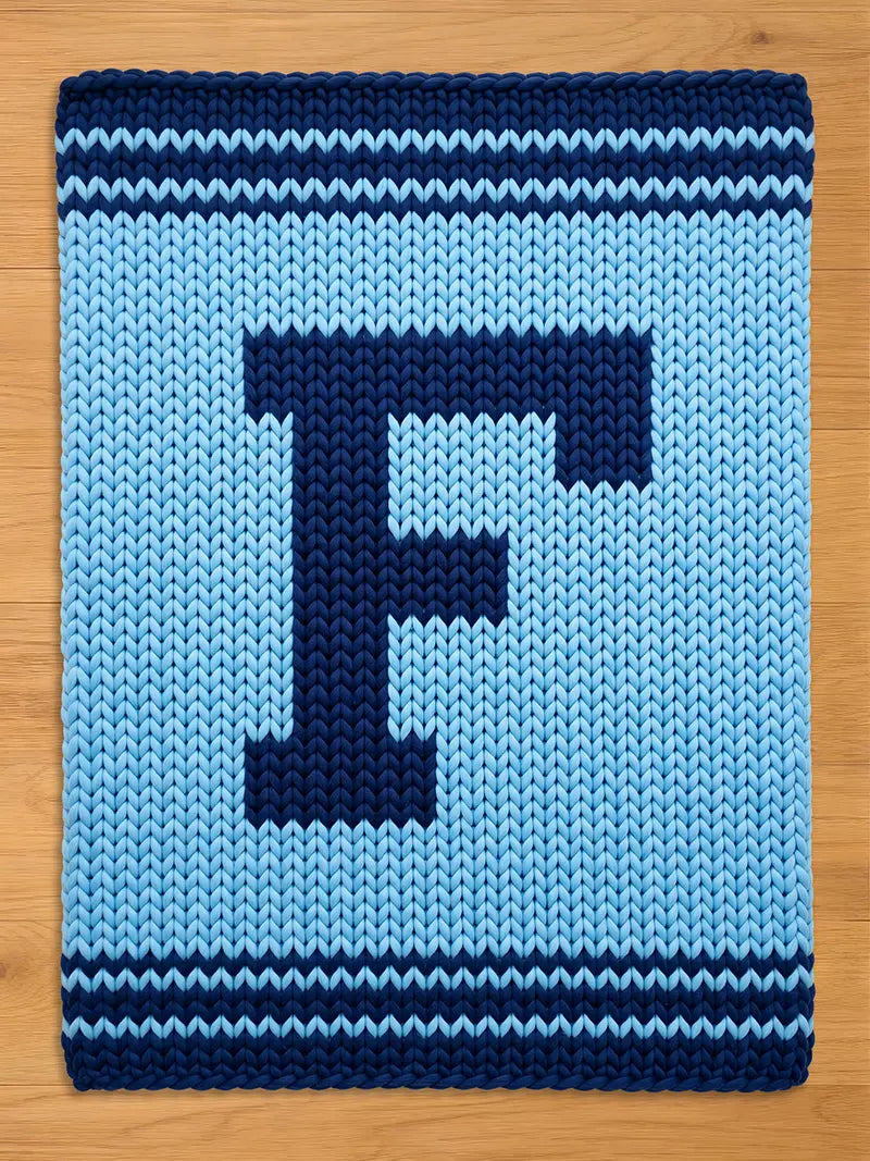 A knitted blanket in shades of blue, featuring a large, dark blue letter "F" in the center. The blanket has a light blue background with dark blue stripes at the top and bottom. The blanket is rectangular.