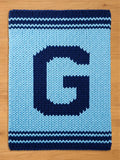 A knitted blanket in light blue with a large, dark blue letter "G" in the center. The blanket has dark blue stripes at the top and bottom edges, creating a contrasting border. The chunky knit texture is visible.