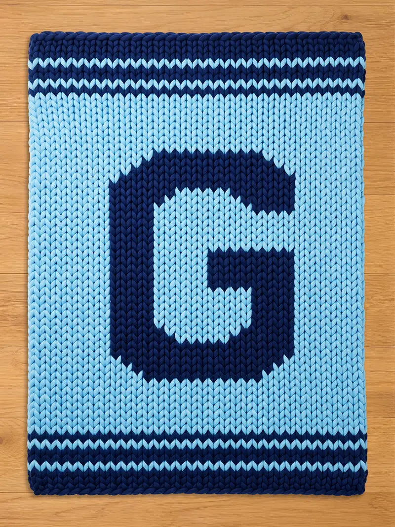 A knitted blanket in light blue with a large, dark blue letter "G" in the center. The blanket has dark blue stripes at the top and bottom edges, creating a contrasting border. The chunky knit texture is visible.