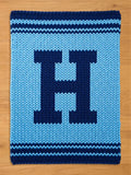 A knitted blanket in shades of blue, featuring a large, dark blue letter "H" in the center. The blanket has a light blue background and dark blue stripes along the top and bottom edges. The blanket is displayed on a wooden surface.