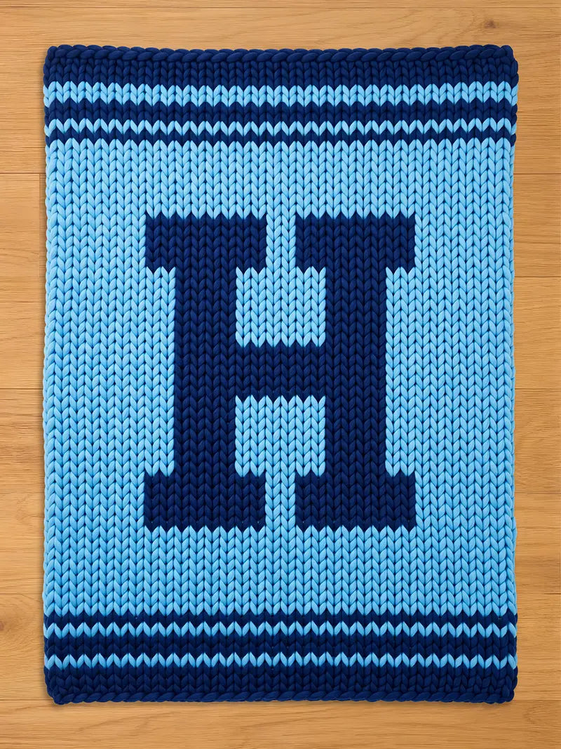 A knitted blanket in shades of blue, featuring a large, dark blue letter "H" in the center. The blanket has a light blue background and dark blue stripes along the top and bottom edges. The blanket is displayed on a wooden surface.