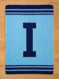 A rectangular, light blue knitted rug with a large, dark blue letter "I" in the center. The rug has dark blue striped borders at the top and bottom, creating a contrasting design. The rug is made of thick yarn.