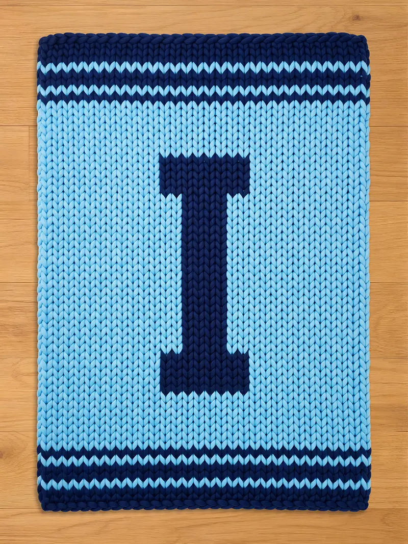 A rectangular, light blue knitted rug with a large, dark blue letter "I" in the center. The rug has dark blue striped borders at the top and bottom, creating a contrasting design. The rug is made of thick yarn.