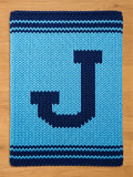 A knitted rectangular blanket in shades of blue. The blanket features a large, dark blue letter "J" in the center. The edges are bordered with dark blue stripes. The background is a lighter shade of blue.