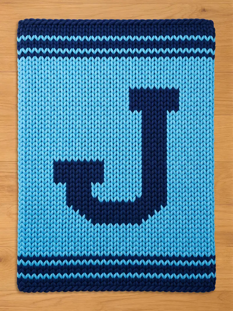 A knitted rectangular blanket in shades of blue. The blanket features a large, dark blue letter "J" in the center. The edges are bordered with dark blue stripes. The background is a lighter shade of blue.