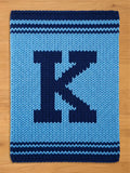 A knitted blanket in shades of blue, featuring a large, dark blue letter "K" in the center. The blanket has a textured appearance with horizontal stripes at the top and bottom. The background is a light blue.