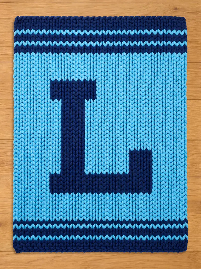 A knitted blue blanket with a large, dark blue letter "L" in the center. The blanket has a light blue background and dark blue stripes along the top and bottom edges. The blanket is made of thick yarn.