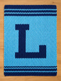 A knitted blue blanket with a large, dark blue letter "L" in the center. The blanket has a light blue background and dark blue stripes along the top and bottom edges. The blanket is made of thick yarn.