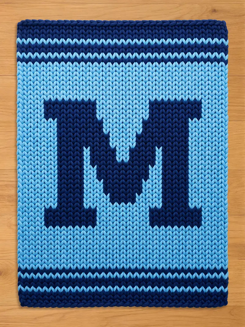 A knitted rectangular blanket in shades of blue. The blanket features a large, dark blue letter "M" in the center, surrounded by a lighter blue background. The top and bottom edges have dark blue stripes.