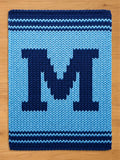 A knitted rectangular blanket in shades of blue. The blanket features a large, dark blue letter "M" in the center, surrounded by a lighter blue background. The top and bottom edges have dark blue stripes.
