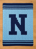 A knitted, rectangular rug in light blue with a large navy blue letter "N" in the center. The rug has navy blue stripes along the top and bottom edges, creating a border. The background is a light wood.