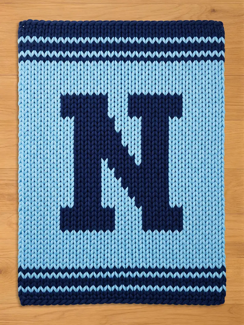 A knitted, rectangular rug in light blue with a large navy blue letter "N" in the center. The rug has navy blue stripes along the top and bottom edges, creating a border. The background is a light wood.