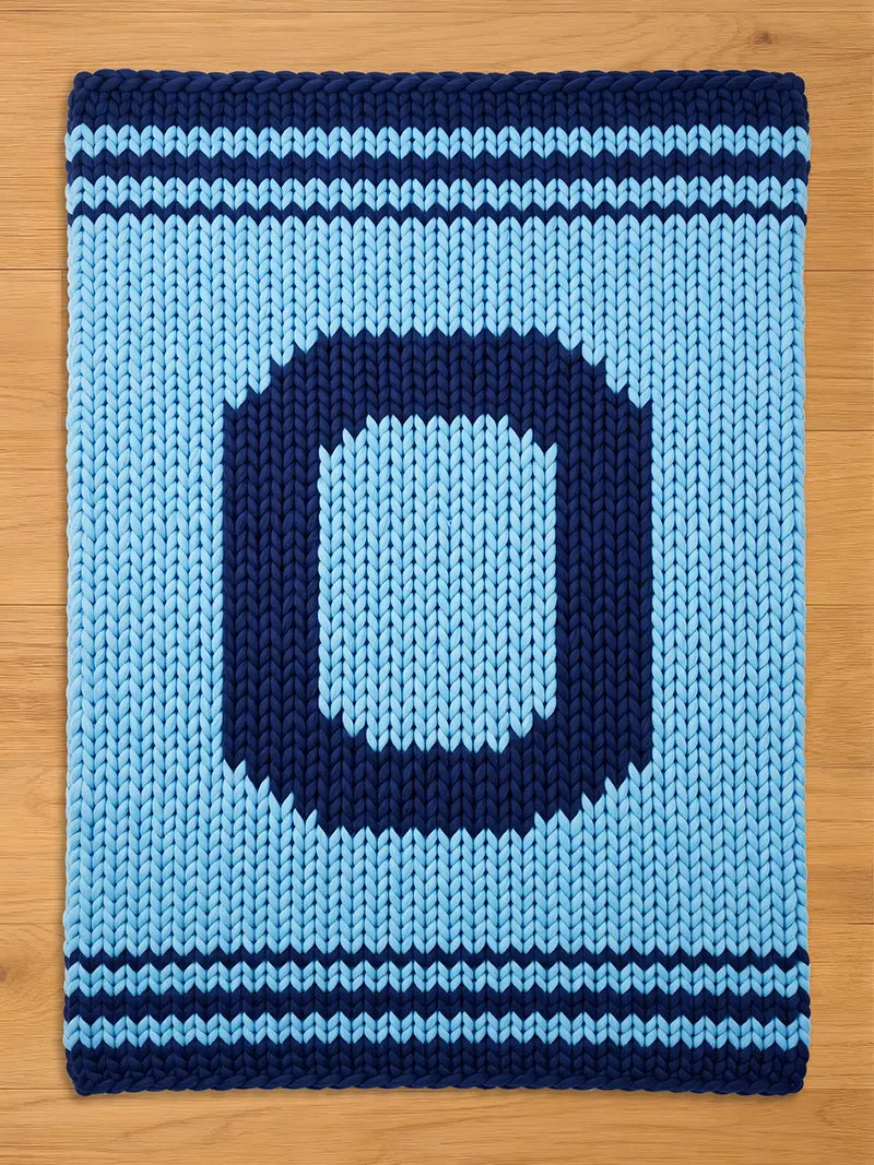 A knitted chunky blanket in shades of blue, featuring a large letter "O" in navy blue against a light blue background. The blanket has navy blue stripes along the top and bottom edges, creating a decorative border. The blanket is made of thick, chunky yarn.