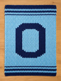 A knitted chunky blanket in shades of blue, featuring a large letter "O" in navy blue against a light blue background. The blanket has navy blue stripes along the top and bottom edges, creating a decorative border. The blanket is made of thick, chunky yarn.