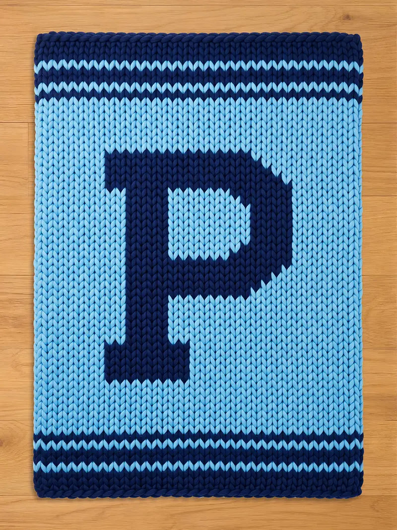 A knitted blanket in light blue with a large navy blue letter "P" in the center. The blanket has navy blue stripes at the top and bottom. The blanket is made of a chunky knit material.