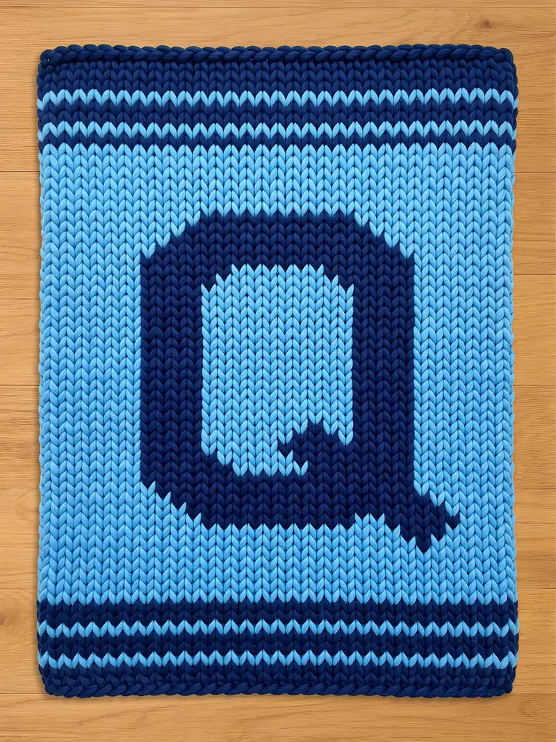 A knitted, rectangular blanket in shades of blue. The blanket features a large, dark blue letter "Q" in the center, surrounded by a lighter blue background. Dark blue stripes border the top and bottom edges. The blanket is made of thick yarn.