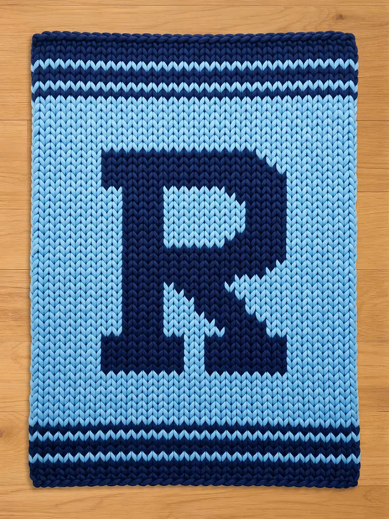A knitted blanket featuring a large letter "R" in navy blue against a light blue background. The blanket has a textured appearance with navy blue stripes along the top and bottom edges. The blanket is displayed on a wooden surface.