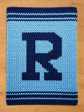 A knitted blanket featuring a large letter "R" in navy blue against a light blue background. The blanket has a textured appearance with navy blue stripes along the top and bottom edges. The blanket is displayed on a wooden surface.
